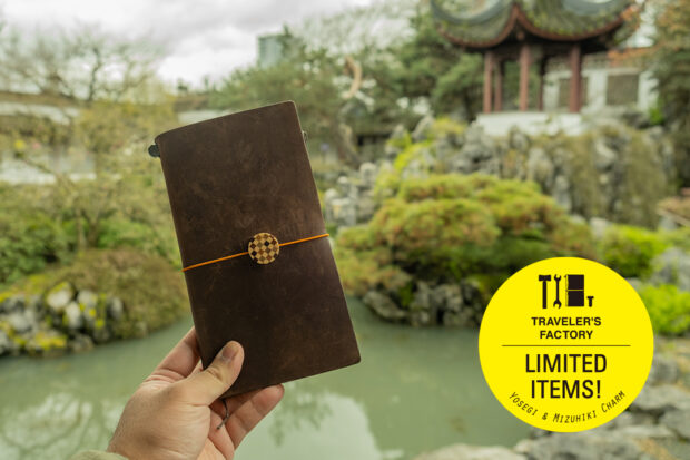 Person holding up a TRAVELER'S notebook with YOSEGI charm against a garden.