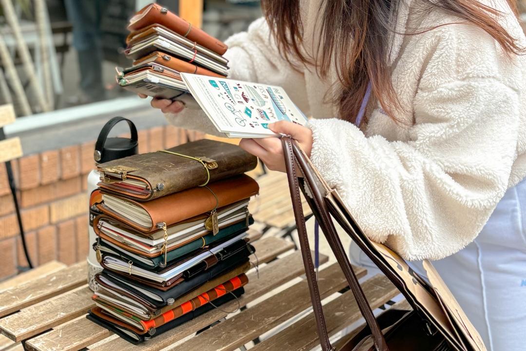 Person stacking TRAVELER'S notebooks on top of each other to create a stack on a wooden table.