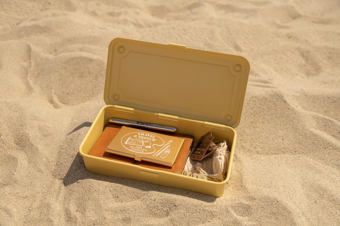 A yellow steel box opens with watercolor supplies in the sand. 