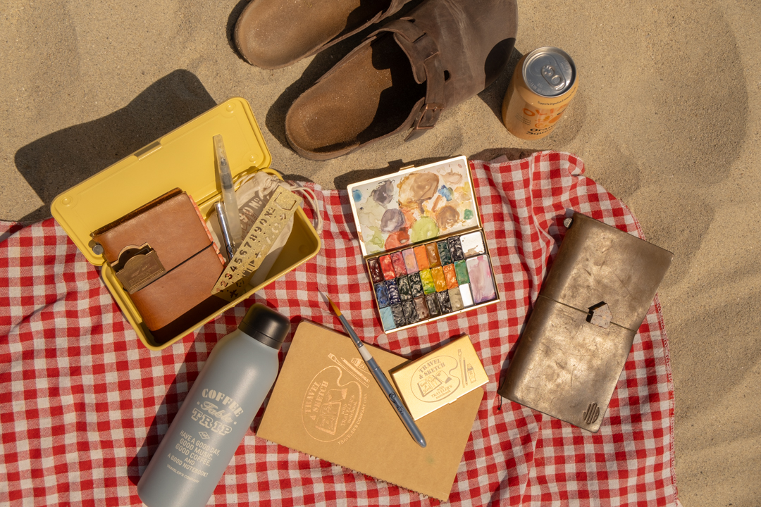 A yellow steel box opens with watercolor supplies, beside a watercolor paint palette, leather notebook, spiral ring notebook, water bottle, soda, and leather sandals on a beach towel in the sand. 