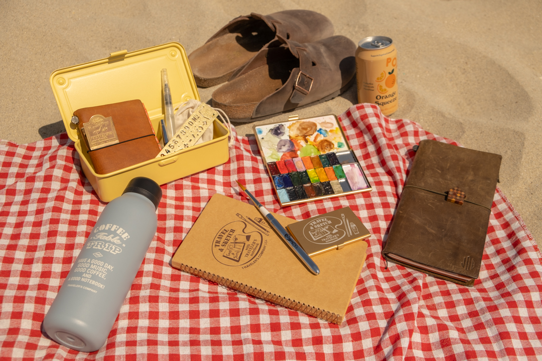 A yellow steel box opens with watercolor supplies, beside a watercolor paint palette, leather notebook, spiral ring notebook, water bottle, soda, and leather sandals on a beach towel in the sand. 