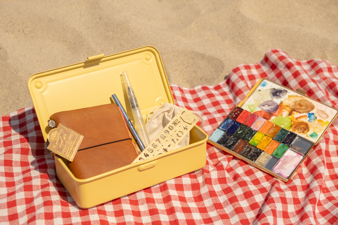 A yellow steel box opens with watercolor supplies, beside a watercolor paint palette on a beach towel. 