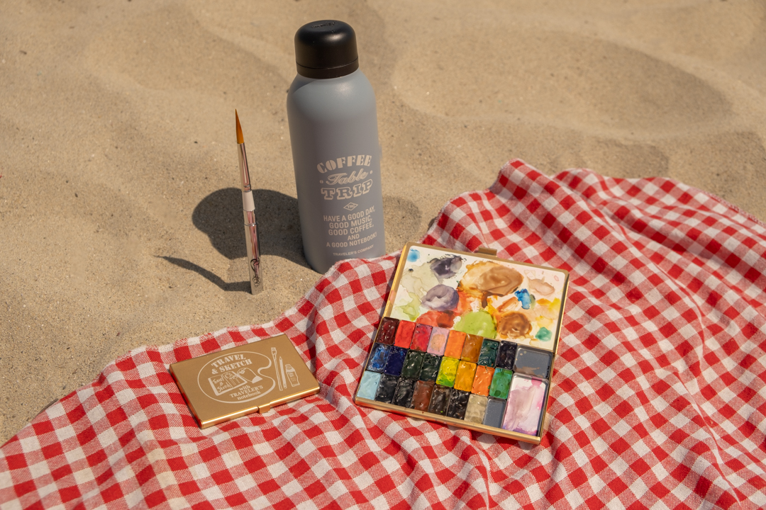 Water bottle, paint brush and two watercolor paint palettes on beach towel on a sandy beach. 