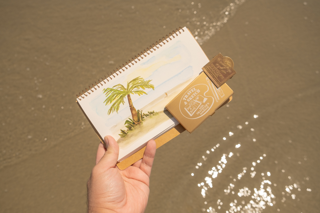 Person holding a watercolor notebook open with sketch of the beach in front of the water at the beach.