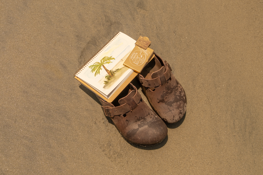 Wet leather sandals on sandy beach with watercolor notebook open with sketch of the beach.