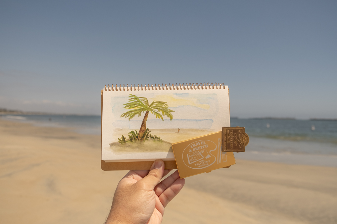 A person holding notebook with a watercolor sketch with a paint palette in front of a beach scene with sand and water. 