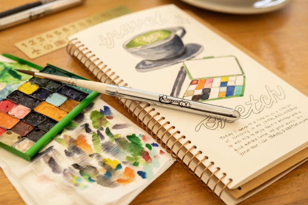 Notebook open with to watercolor sketch of cup and paint palette beside a matcha latte and pens and paint brushes.