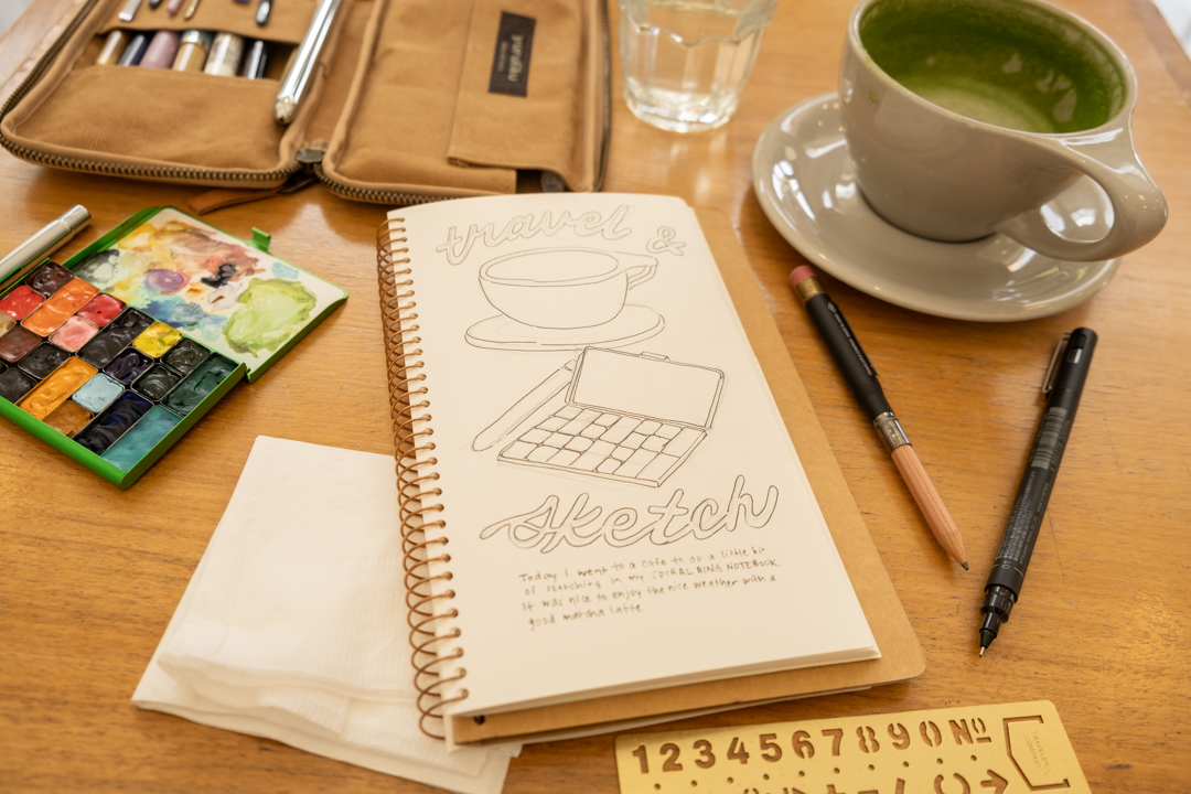 Notebook open with sketch of cup and paint palette beside a matcha latte and pens and paint brushes.