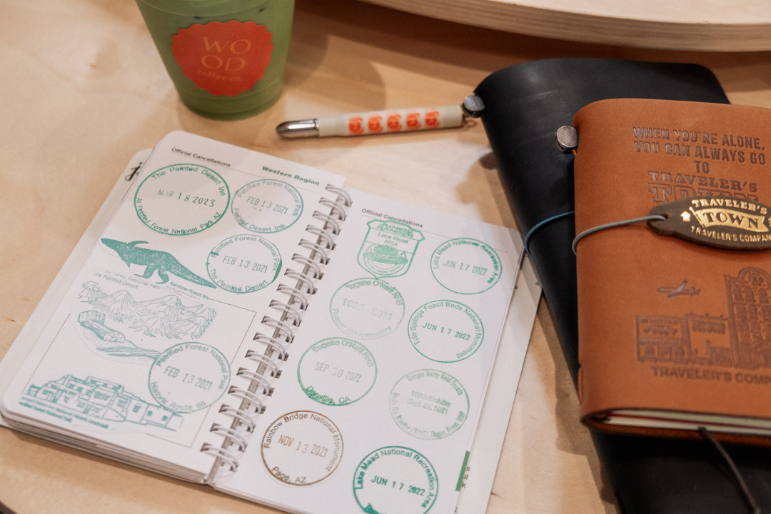 National Park stamps on little sheets of paper on a wooden table beside leather journals, coffe and a pen. 