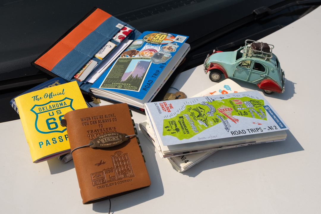 A TRAVELER'S notebook open to show a notebook insert decorated with Road Trip stickers beside other notebooks on top of a car hood.