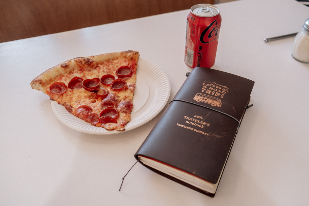 TRAVELER'S notebook next to a slice of pizza and a can of soda.