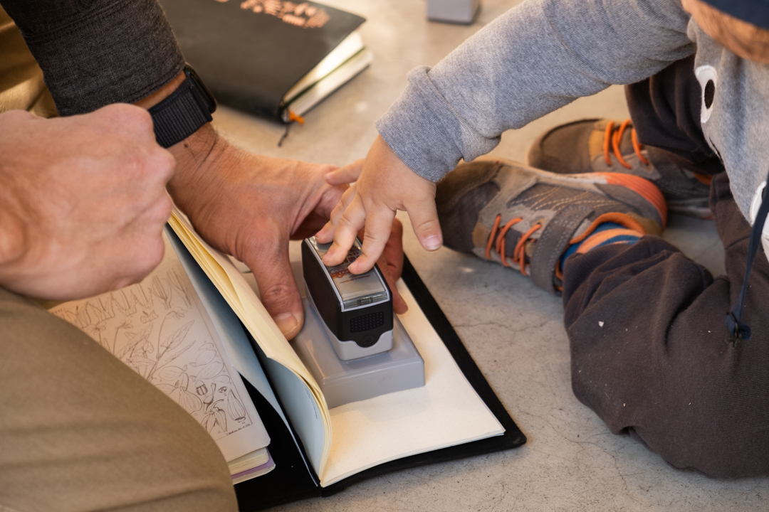 A toddler using an ink stamp to stamp a notebook with the help of their father.