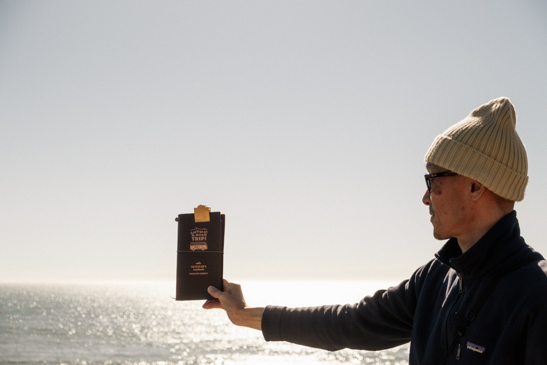 Man holding a TRAVELER'S notebook in front of the ocean.