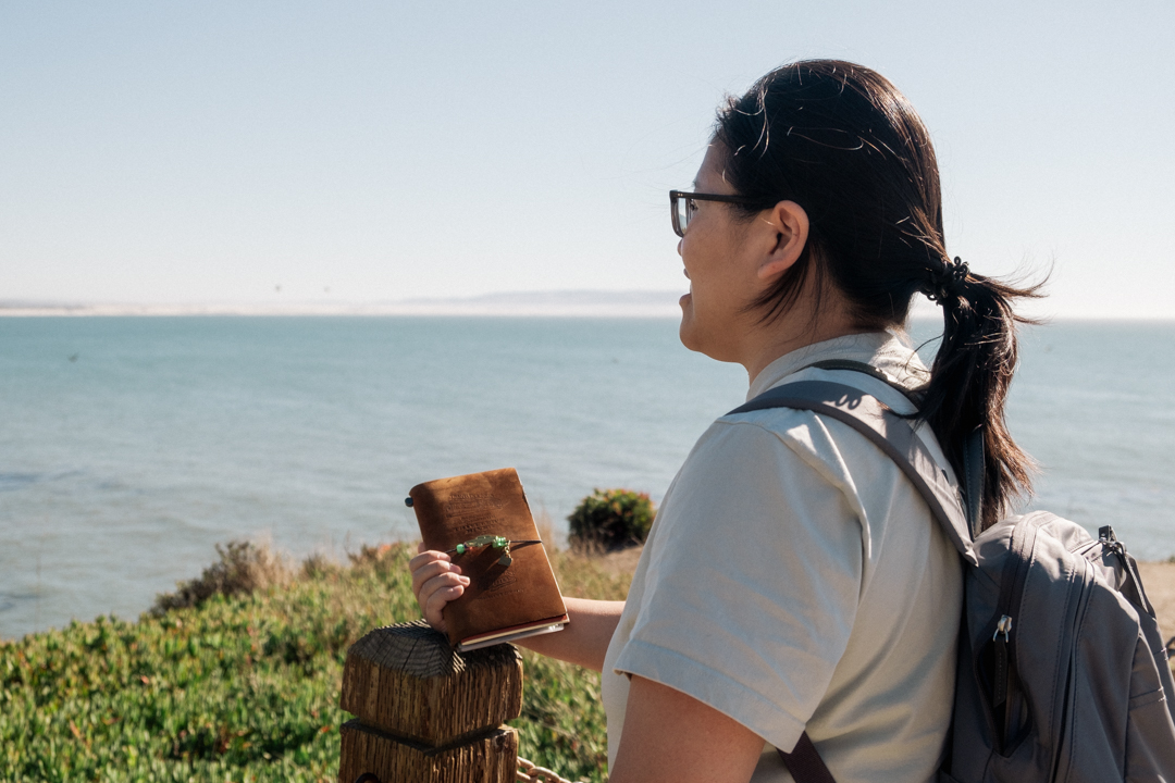 Woman holding a TRAVELER'S notebook while admiring the view of the oceanside cliff.