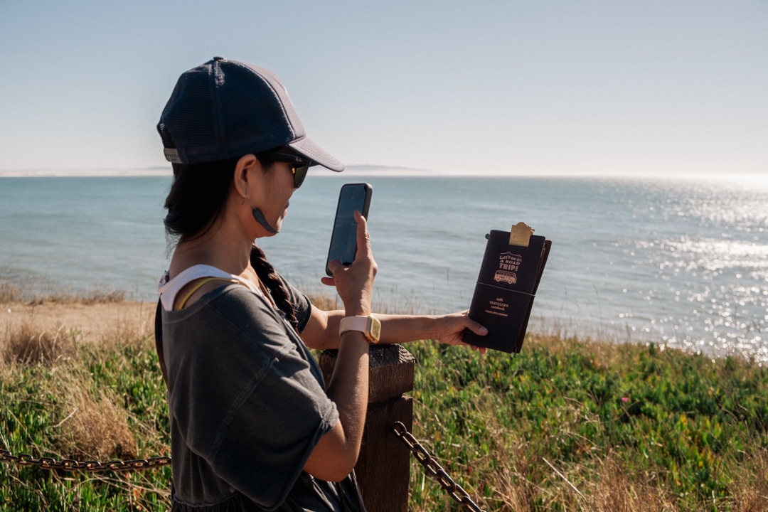 Woman holding a TRAVELER'S notebook in front of an oceanside cliff.