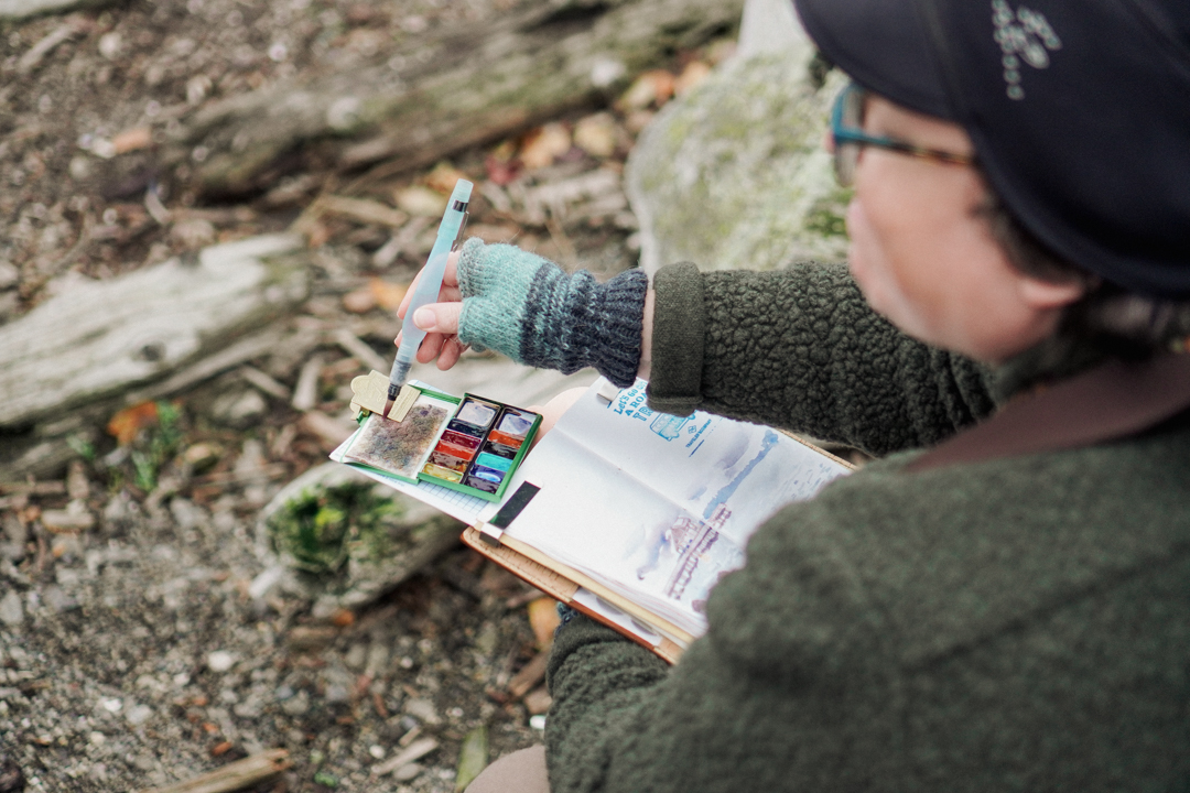 Person painting in their TRAVELER'S notebook while using their Art Toolkit paint palette and brush.