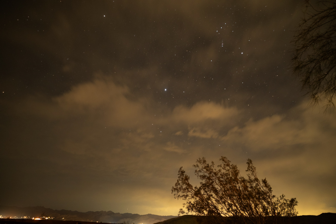 Photo of the night sky that is partially cloudy with some stars and a tree on the side.