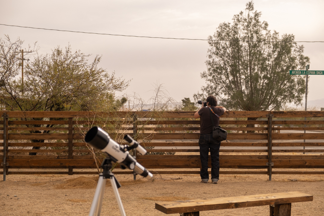 Man taking photos by a fence facing trees and the desert.