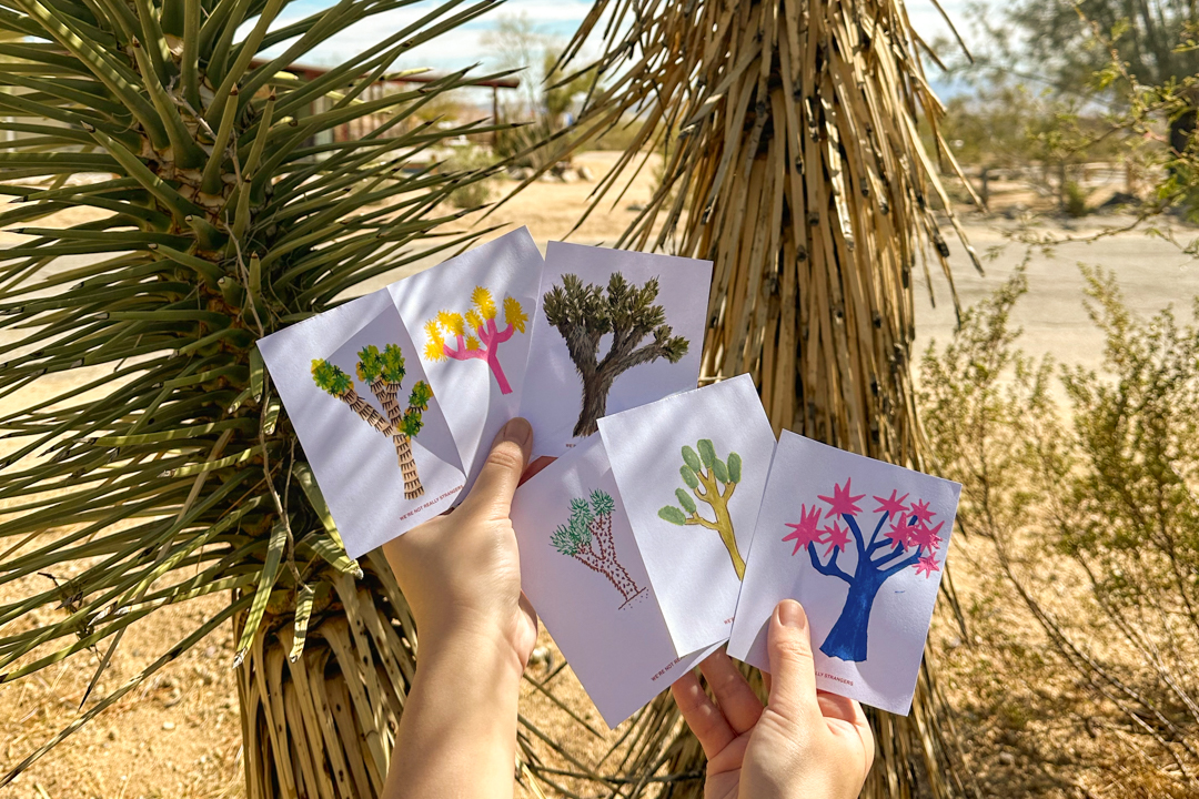 Person holding 6 different illustrations of Joshua Trees in front of a Joshua Tree.