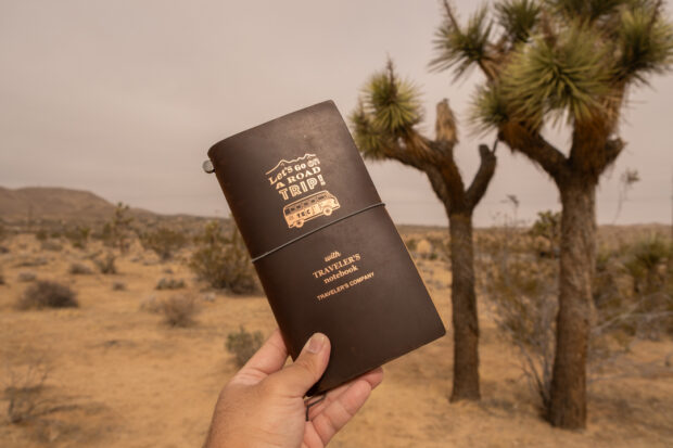 A person holding a Road Trip TRAVELER'S notebook in front of large Joshua Trees in the Joshua Tree National Park in California.