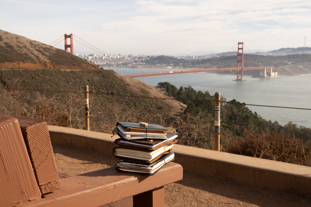 TRAVELER'S notebooks stacked on a bench in front of a view of the Golden Gate Bridge in San Francisco.
