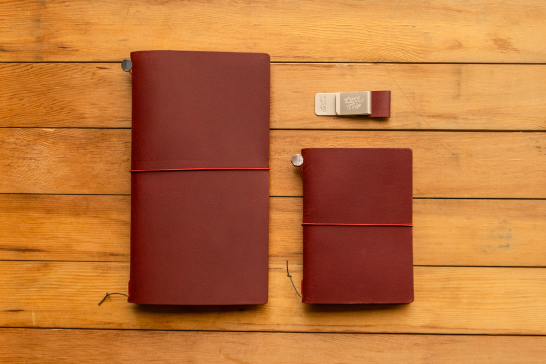 The LOVE AND TRIP red TRAVELER'S notebook in regular and passport size beside a pen holder.