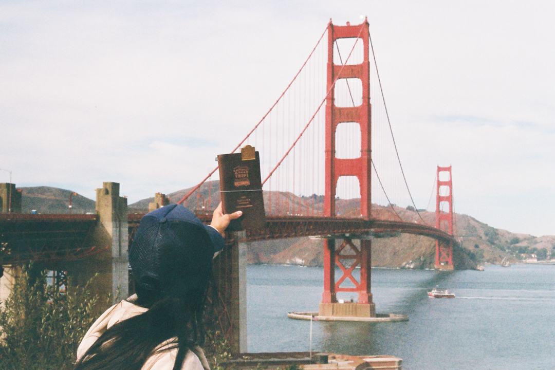 A person holding a TRAVELER'S notebook in front of the Golden Gate Bridge.
