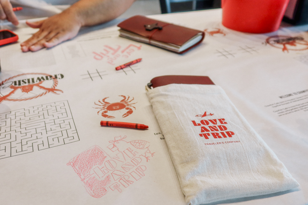 Crayon doodles on a white paper table cloth beside a TRAVELER'S notebook in a cloth case. 