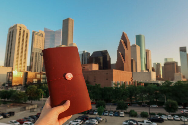 A person holding a red TRAVELER'S notebook in front of the Houston, Texas skyline.