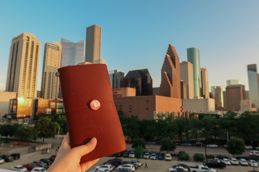 A person holding a red TRAVELER'S notebook in front of the Houston, Texas skyline.