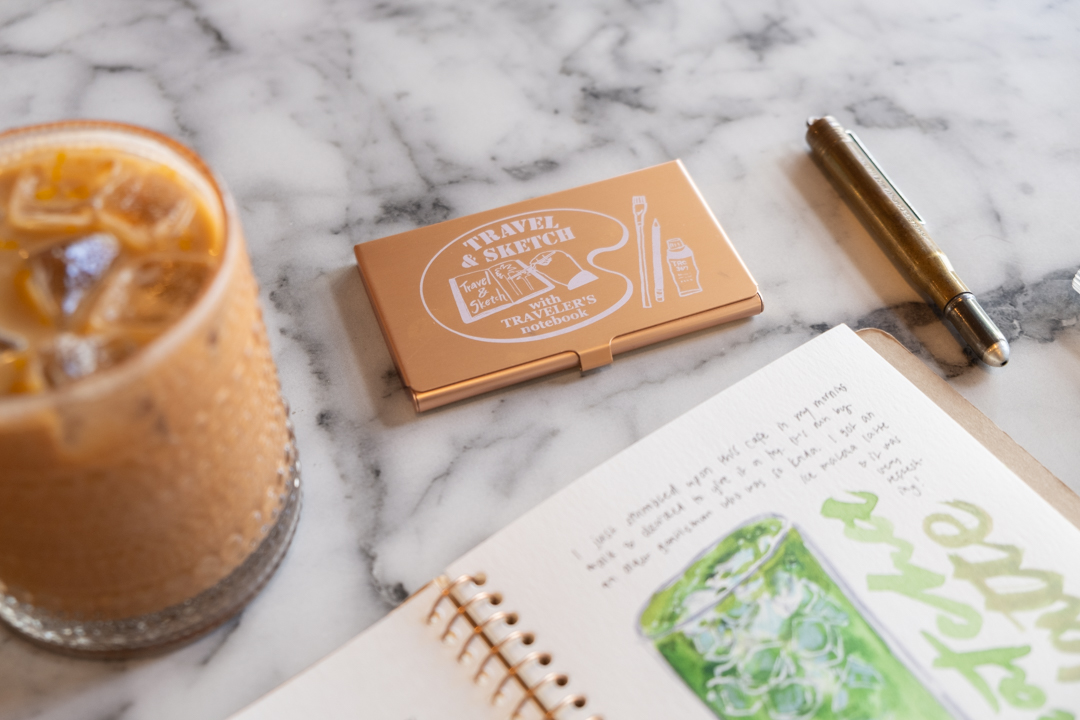 A close up look at the TRC USA x Art Toolkit Pocket Palette next to a glass of iced coffee. 