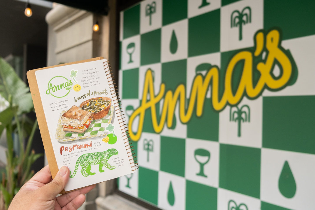 A hand holding up a TRAVEL & SKETCH SPIRAL RING NOTEBOOK with an illustration of a sandwich and store logo next to the green checkered interior wall of Anna's in Long Beach.