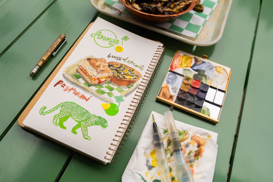 A drawing of a sandwich and a bowl of brussels sprouts on a TRAVEL & SKETCH SPIRAL RING NOTEBOOK next to a TRC USA x Art Toolkit Pocket Palette.