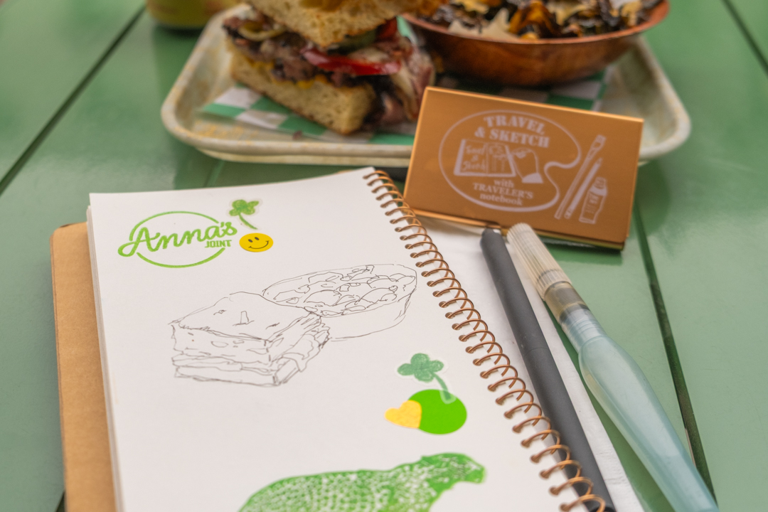 A half-finished drawing of a sandwich and a bowl of brussels sprouts on a TRAVEL & SKETCH SPIRAL RING NOTEBOOK next to a TRC USA x Art Toolkit Pocket Palette.