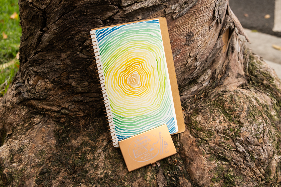 A TRAVEL & SKETCH SPIRAL RING NOTEBOOK with a tree ring painting leaning against a tree trunk with a TRC USA x Art Toolkit Pocket Palette. 