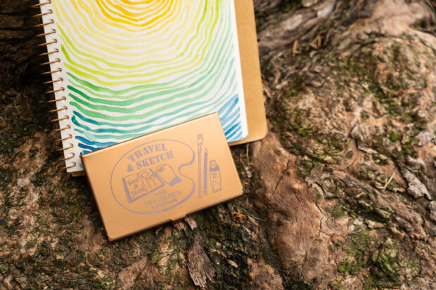 A TRC USA Limited Art Toolkit Pocket Palette leaning against a painting of colorful tree rings against a tree trunk.