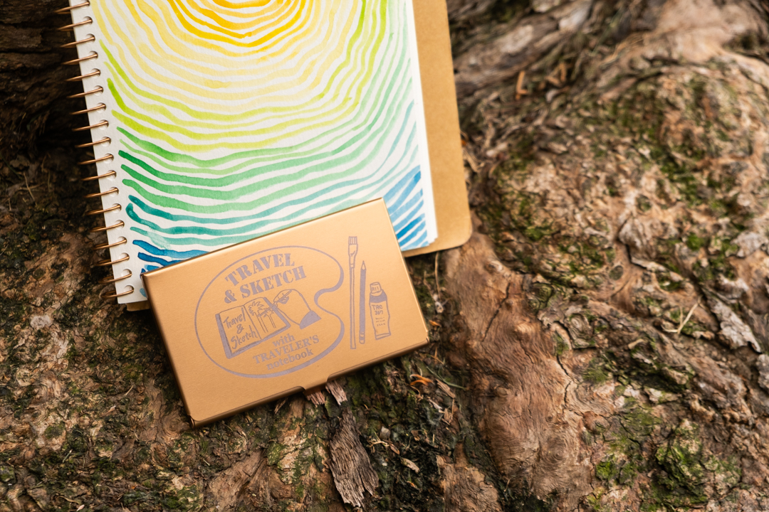 A TRC USA Limited Art Toolkit Pocket Palette leaning against a painting of colorful tree rings against a tree trunk.