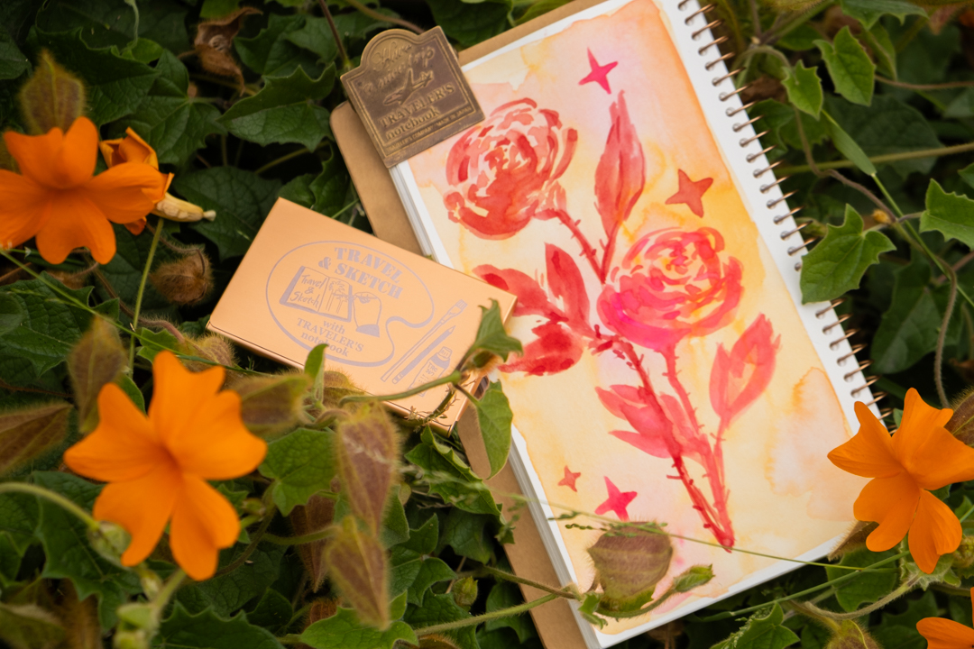 A painting of red roses on a TRAVEL & SKETCH SPIRAL RING NOTEBOOK in a flower bush next to a TRC USA x Art Toolkit Pocket Palette. 