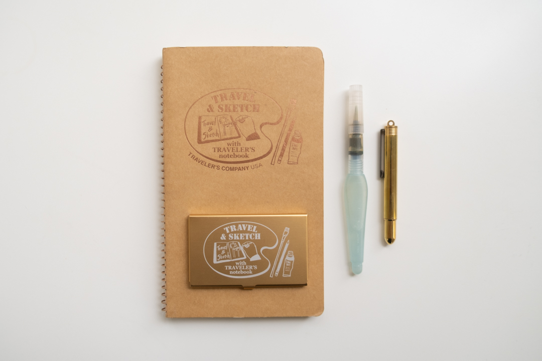 A TRAVEL & SKETCH SPIRAL RING NOTEBOOK (A5 Slim), a TRC USA x Art Toolkit Pocket Palette, a Pentel Mini Waterbrush, and a BRASS PENCIL on a white surface. 