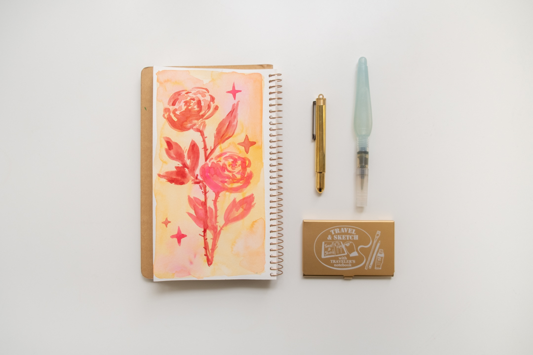 A painting of red roses on a TRAVEL & SKETCH SPIRAL RING NOTEBOOK next to a BRASS PENCIL, Mini waterbrush, and a TRC USA x Art Toolkit Pocket Palette.