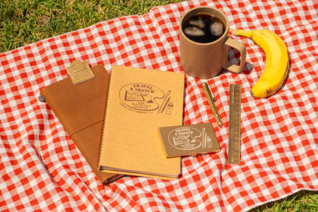 A TRAVELER'S notebook, a TRAVEL & SKETCH SPIRAL RING NOTEBOOK, and an collaboration Art Toolkit Palette together on a picnic blanket.
