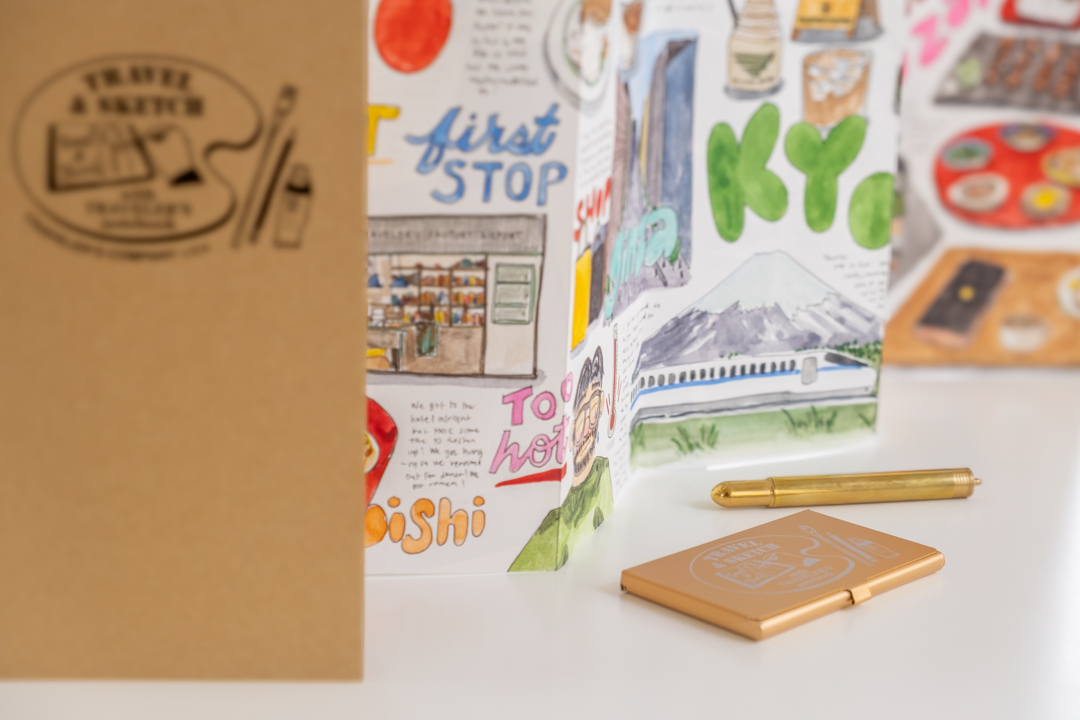 An Accordion Fold Paper TRAVEL & SKETCH filled with drawings, next to a TRAVEL & SKETCH Pocket Palette and BRASS BALLPOINT PEN.