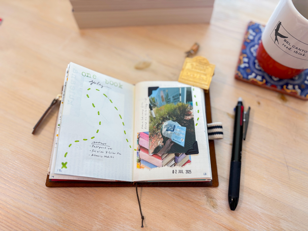 A brown passport TRAVELER'S notebook open to a travel journal spread beside a mug, a pen, and books on a wooden table.