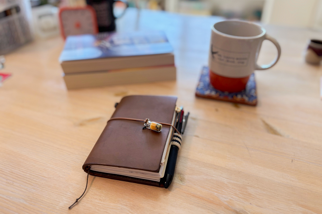 A brown passport TRAVELER'S notebook on a wooden table beside a mug and books.