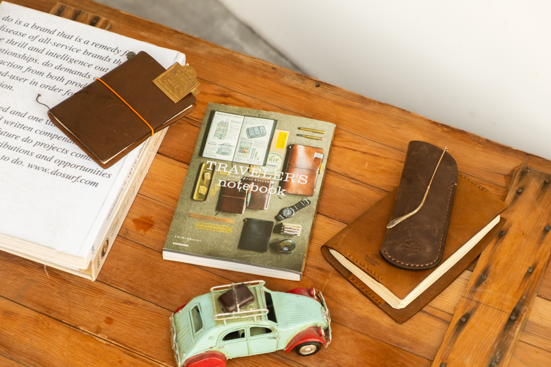 The TRAVELER'S GUIDE book next to other books, a TRAVELER'S notebook in brown, a toy car and leather book cover with a leather glasses case in brown on top of it.