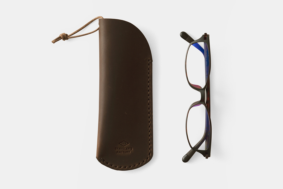 A leather glasses case beside narrow frame black glasses on a white background.