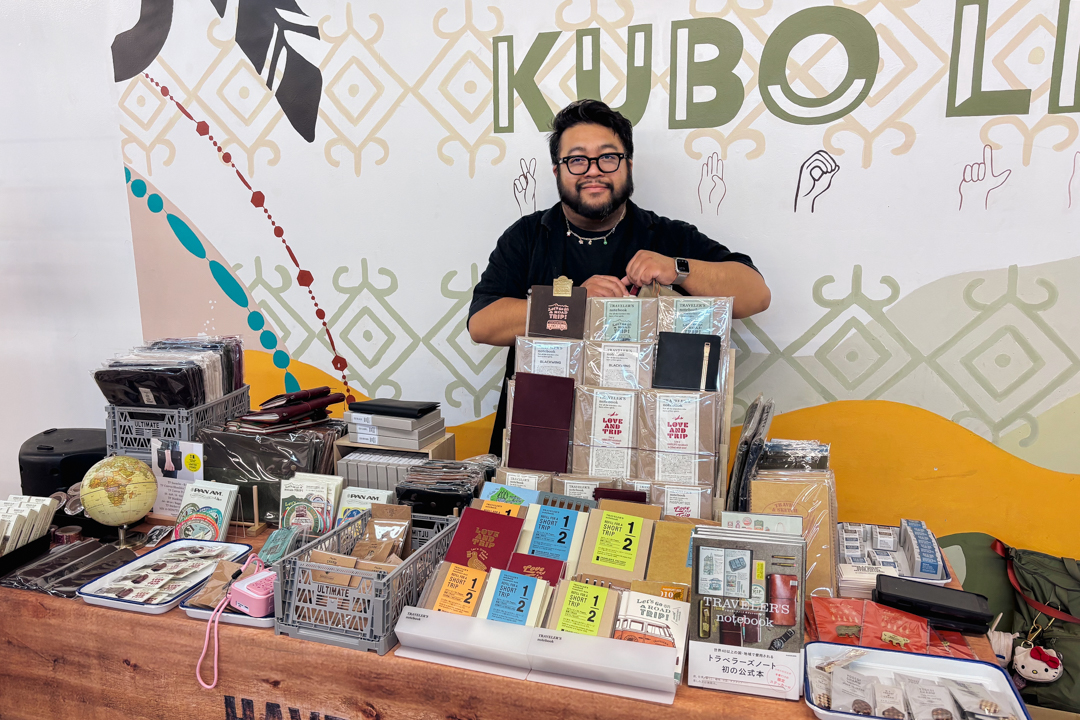 TRC USA staff standing behind the TRC USA booth at KUBO LB, featuring books, paper goods, and stationery on the vendor table. The atmosphere is lively and engaging.