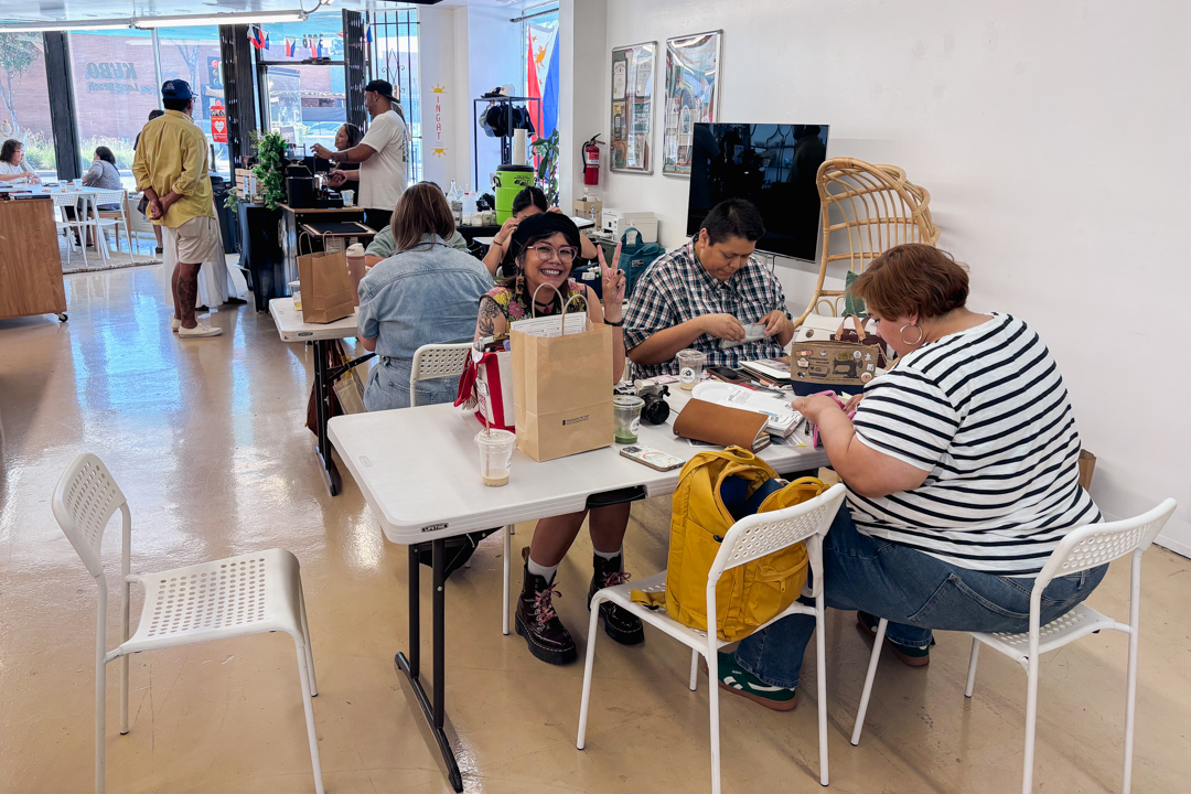 People sit at tables inside KUBO LB with white walls and ample light. One person smiles and makes a peace sign, creating a friendly, relaxed atmosphere.