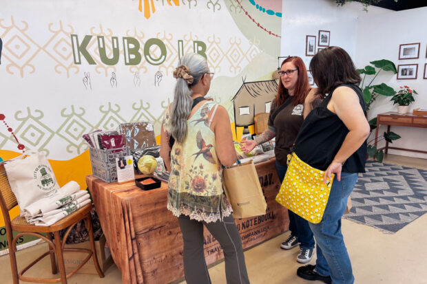 Two women shop at the TRC USA booth at Kubo LB, speaking to another customer. The booth displays stationery, and TRC items, creating a vibrant, friendly atmosphere.