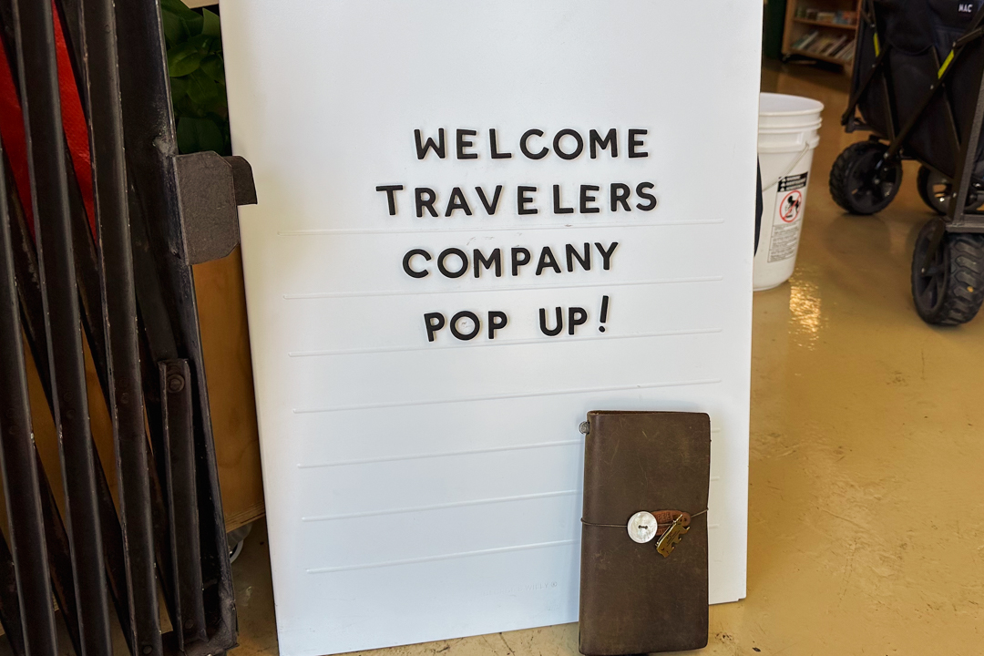 A floor sign displaying "Welcome TRAVELER'S COMPANY POP UP" beside a regular size TRAVELER'S notebook.